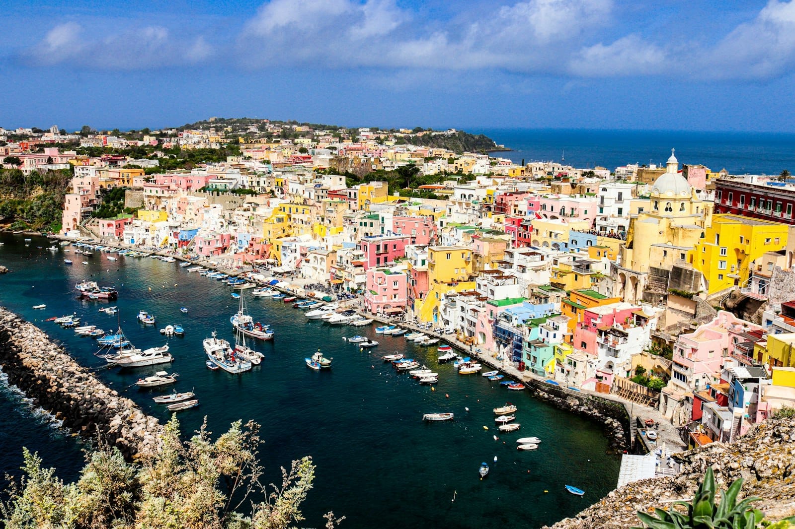 Panoramica delle colorate case pastello del porto dell'isola di Procida e delle barche da pesca alla luce del giorno, con il mare e il cielo blu sullo sfondo.