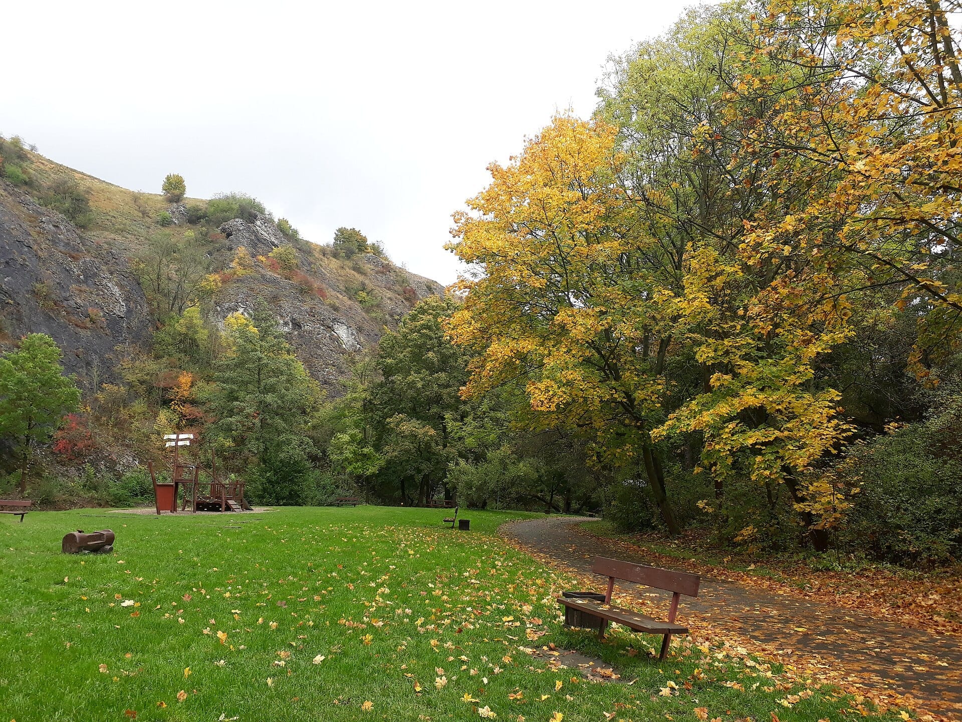 Herbstszene im Prokop-Tal mit einem gewundenen Pfad, Holzbänken, grünem Rasen, bunten Bäumen und felsigen Hügeln im Hintergrund.