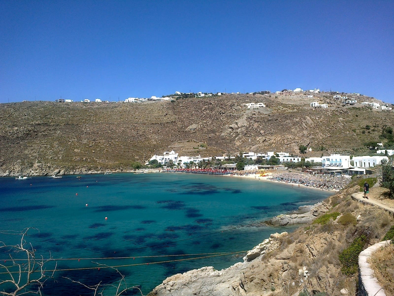 Playa de Psarou en Mykonos con aguas turquesas, arena clara, tumbonas y edificios blancos frente a una escarpada ladera bajo cielos azules despejados.