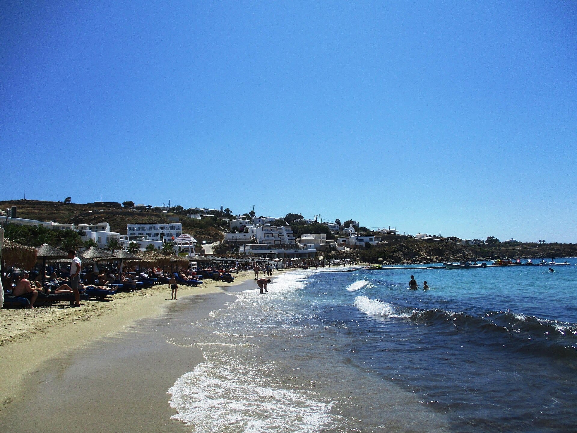 Día soleado en la playa de Platis Gialos en Mykonos, con personas relajándose en tumbonas, mar azul cristalino y edificios encalados en la ladera.