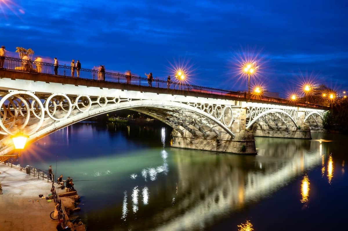 สะพาน Puente de Triana ที่สว่างไสวในยามค่ำคืน พร้อมนักเดินเท้าบนสะพานและภาพสะท้อนของแสงไฟถนนบนแม่น้ำ Guadalquivir ที่สงบด้านล่าง