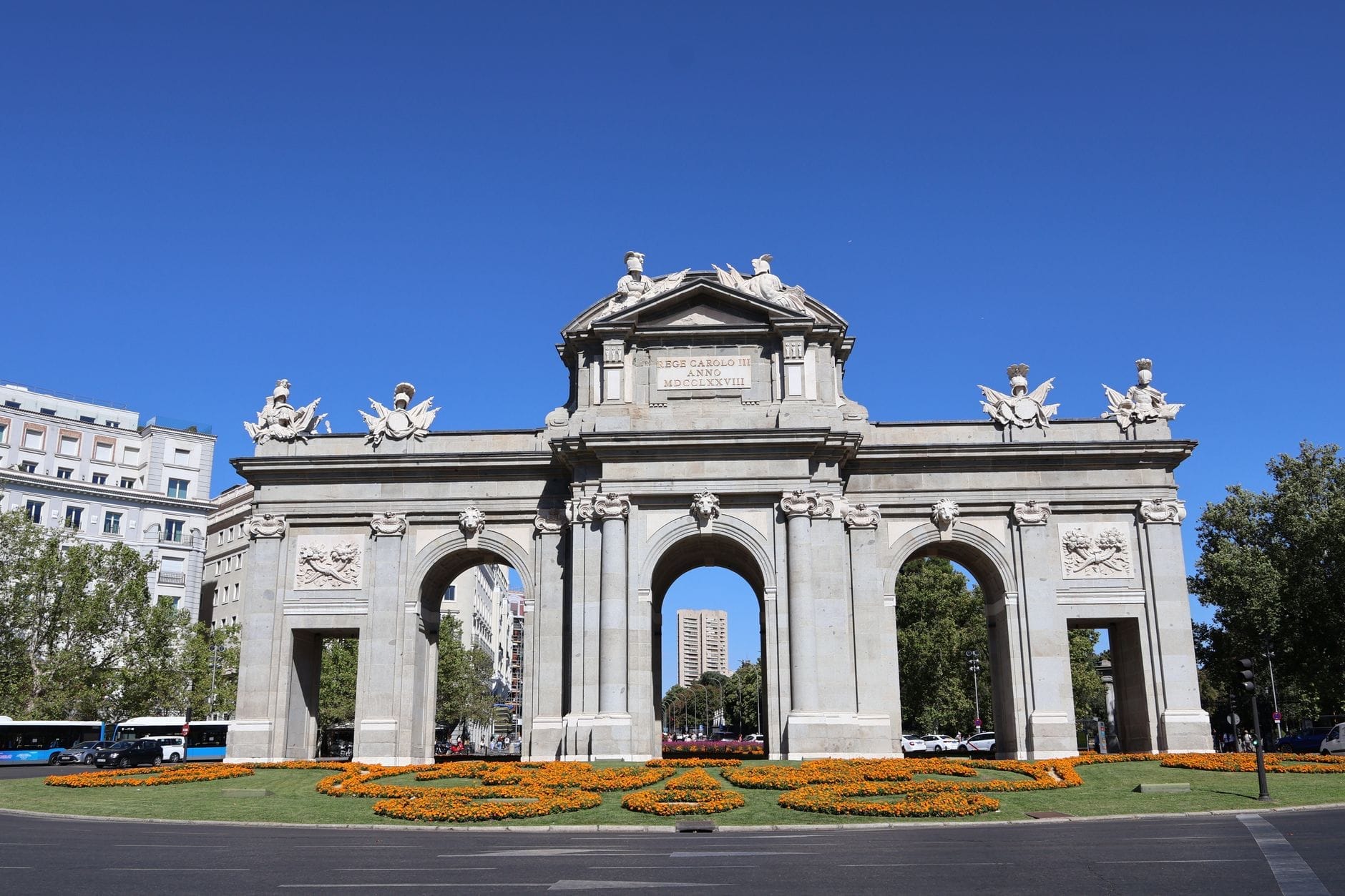 Vista nitida e luminosa della Puerta de Alcalá a Madrid con cielo azzurro, sculture elaborate e aiuole di fiori arancioni in primo piano, incorniciata dagli edifici circostanti.