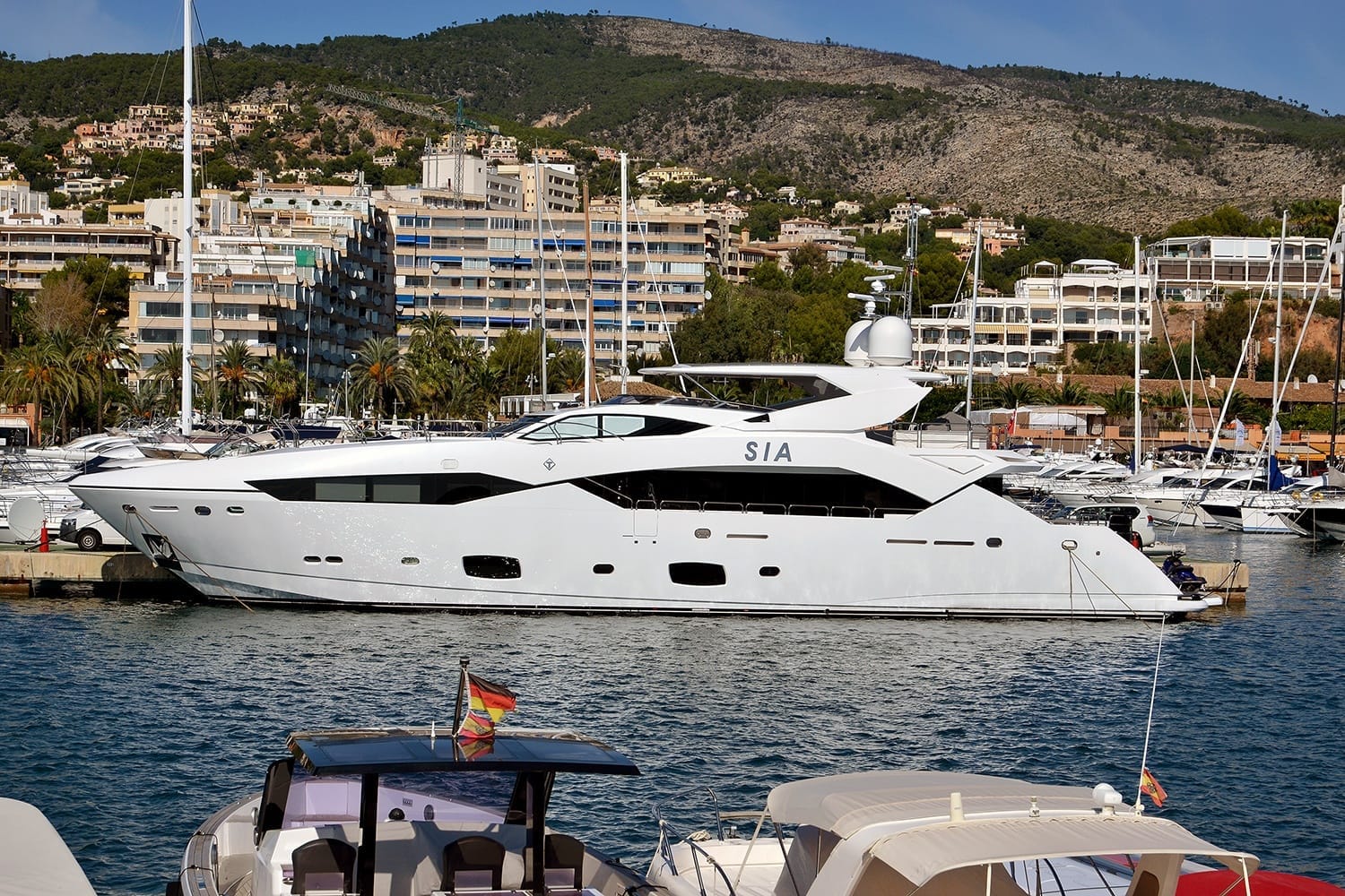 Grande yacht bianco ormeggiato al porto di Puerto Portals con moderni edifici residenziali, colline verdi e palme sullo sfondo alla luce del giorno.