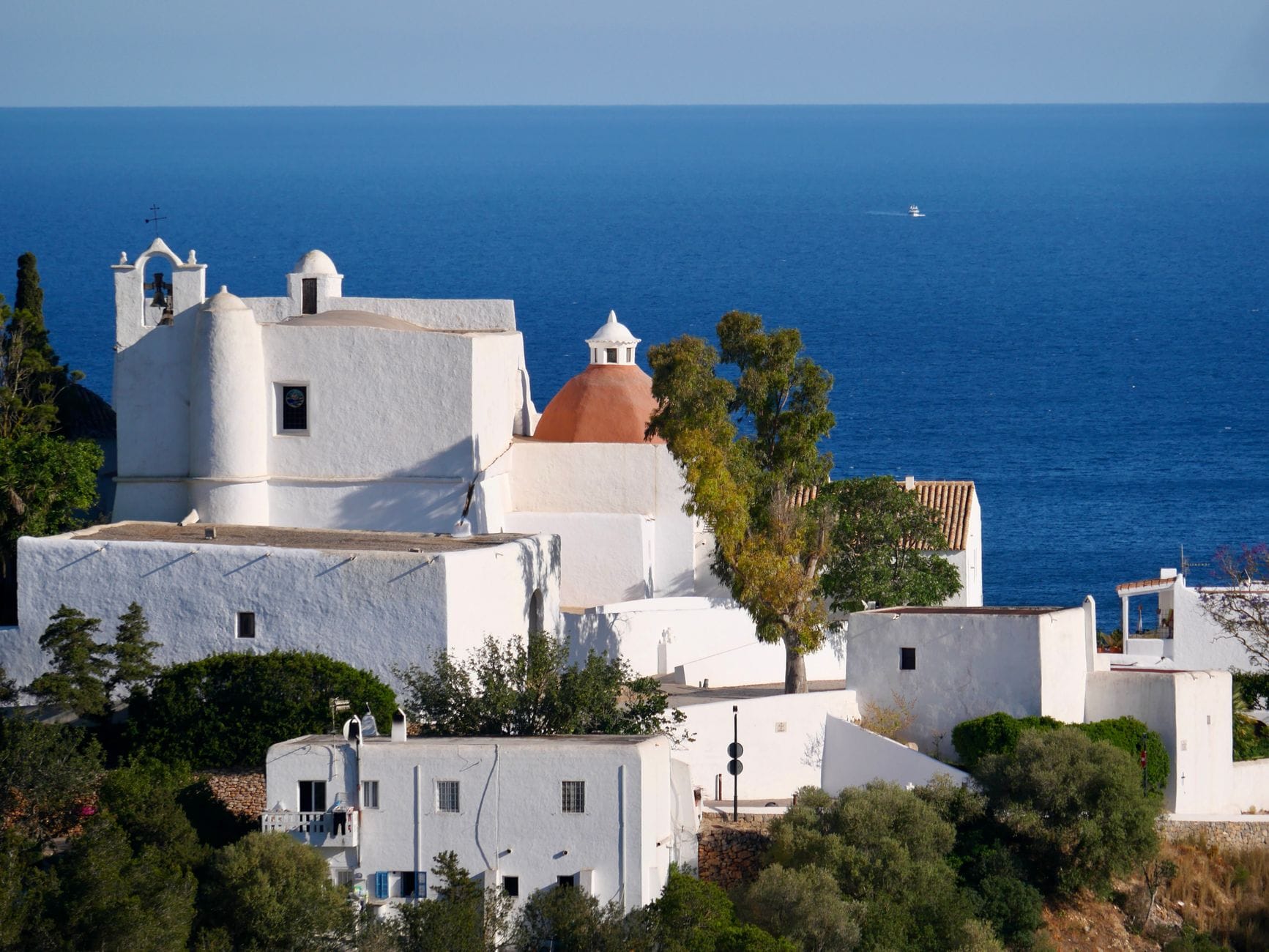 La chiesa imbiancata di Puig de Missa arroccata su una collina con vista sul mare, circondata da alberi e edifici storici a Santa Eulalia, Ibiza.