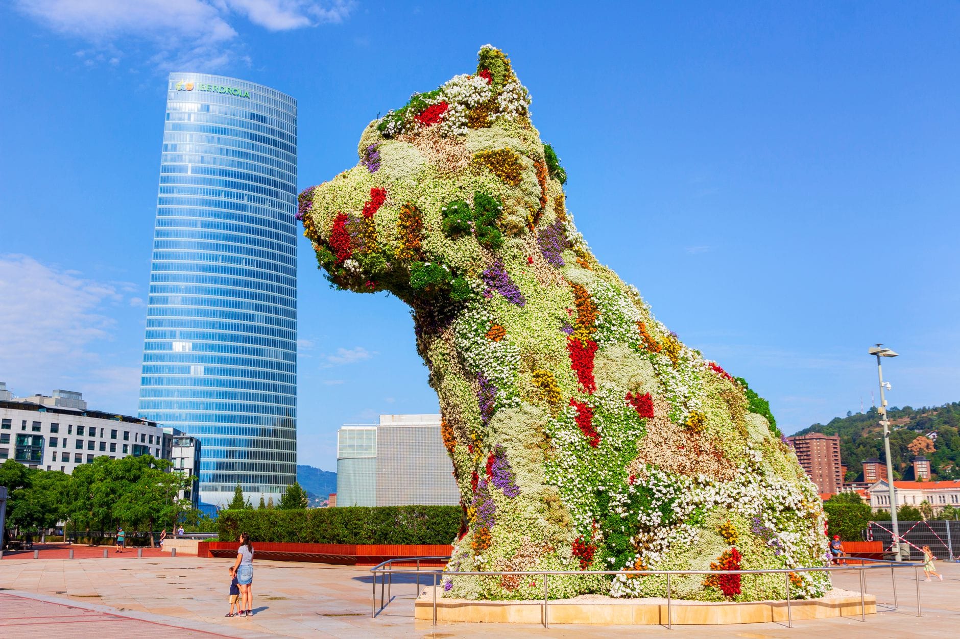 Jeff Koons' Puppy sculpture covered in colorful flowers, prominently displayed in front of the Guggenheim Museum Bilbao on a sunny day with modern buildings in the background.