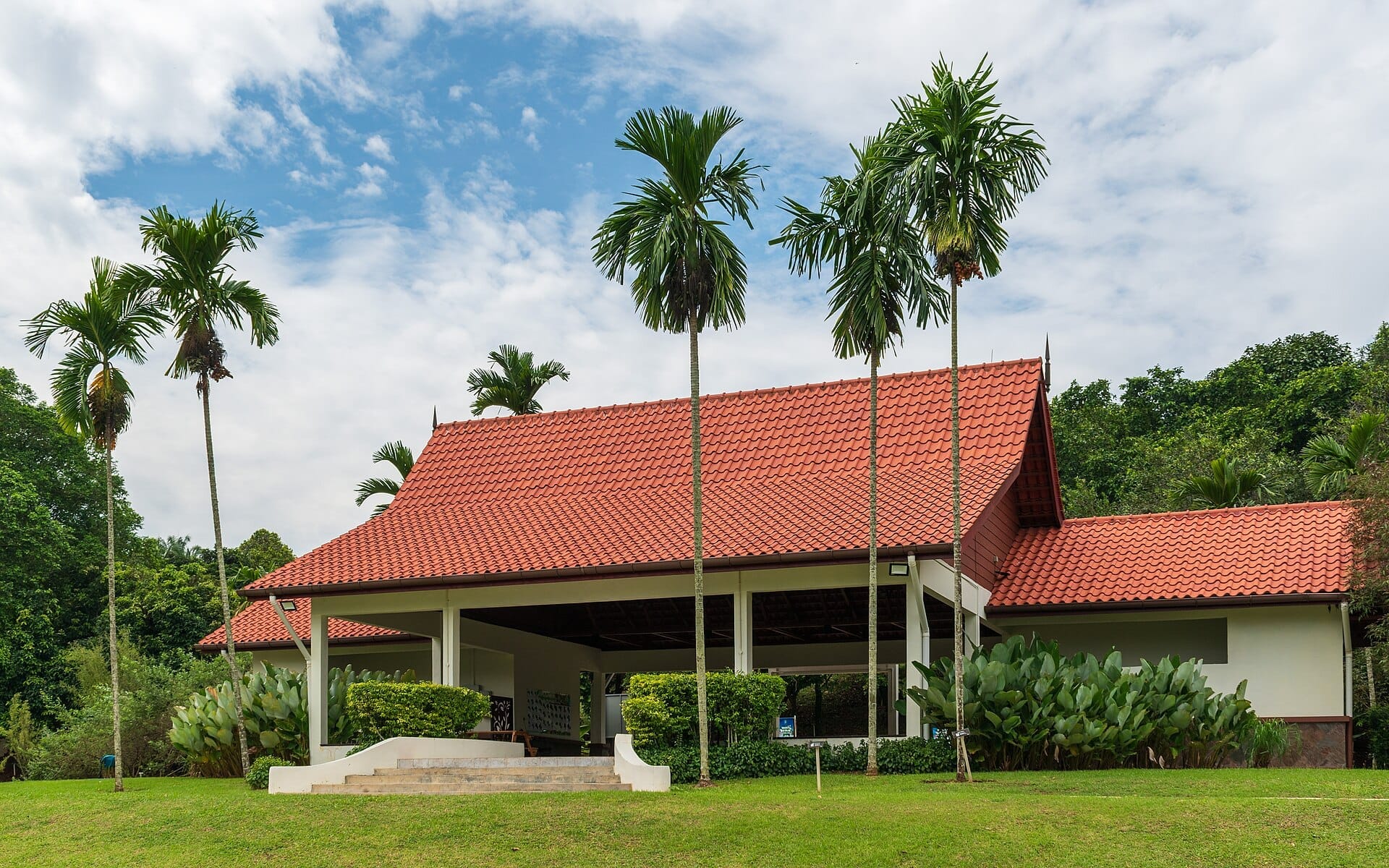 Red-roofed Ethnobotany pavilion surrounded by lush tropical plants and palm trees at Putrajaya Botanical Garden under a partly cloudy sky.