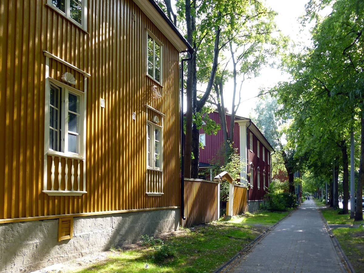 Casas de madeira tradicionais em Puu-Käpylä, Helsinque, com fachadas amarelas e vermelhas ao longo de um caminho pedonal arborizado num dia ensolarado.