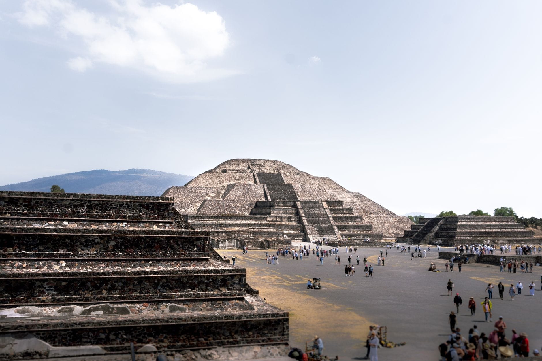 Vista ampia della Piramide della Luna a Teotihuacán con visitatori in primo piano e le antiche strutture circostanti sotto un cielo luminoso.