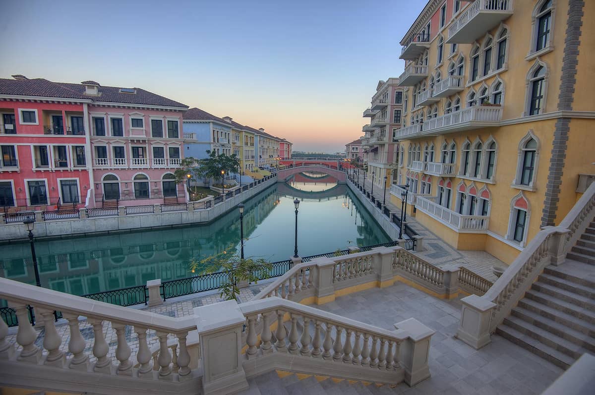 Elegante Pastellgebäude entlang eines ruhigen Kanals mit Bogenbrücken im Qanat Quartier auf The Pearl-Qatar, bei Sonnenuntergang von einer breiten Treppe aus fotografiert.