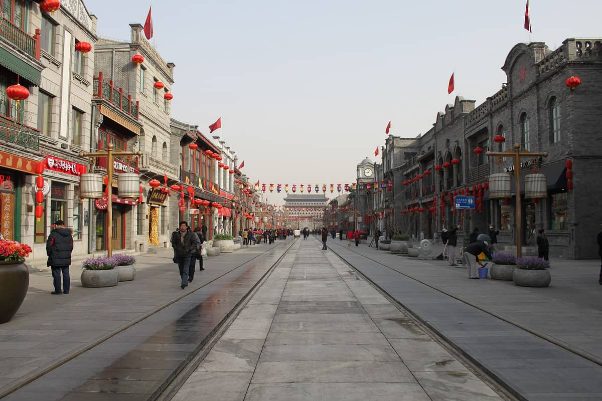 Weiter Blick die Qianmen-Straße in Peking hinunter, gesäumt von historisch gestalteten Gebäuden und roten Laternen, mit Passanten und dem Zhengyangmen-Tor am fernen Ende.