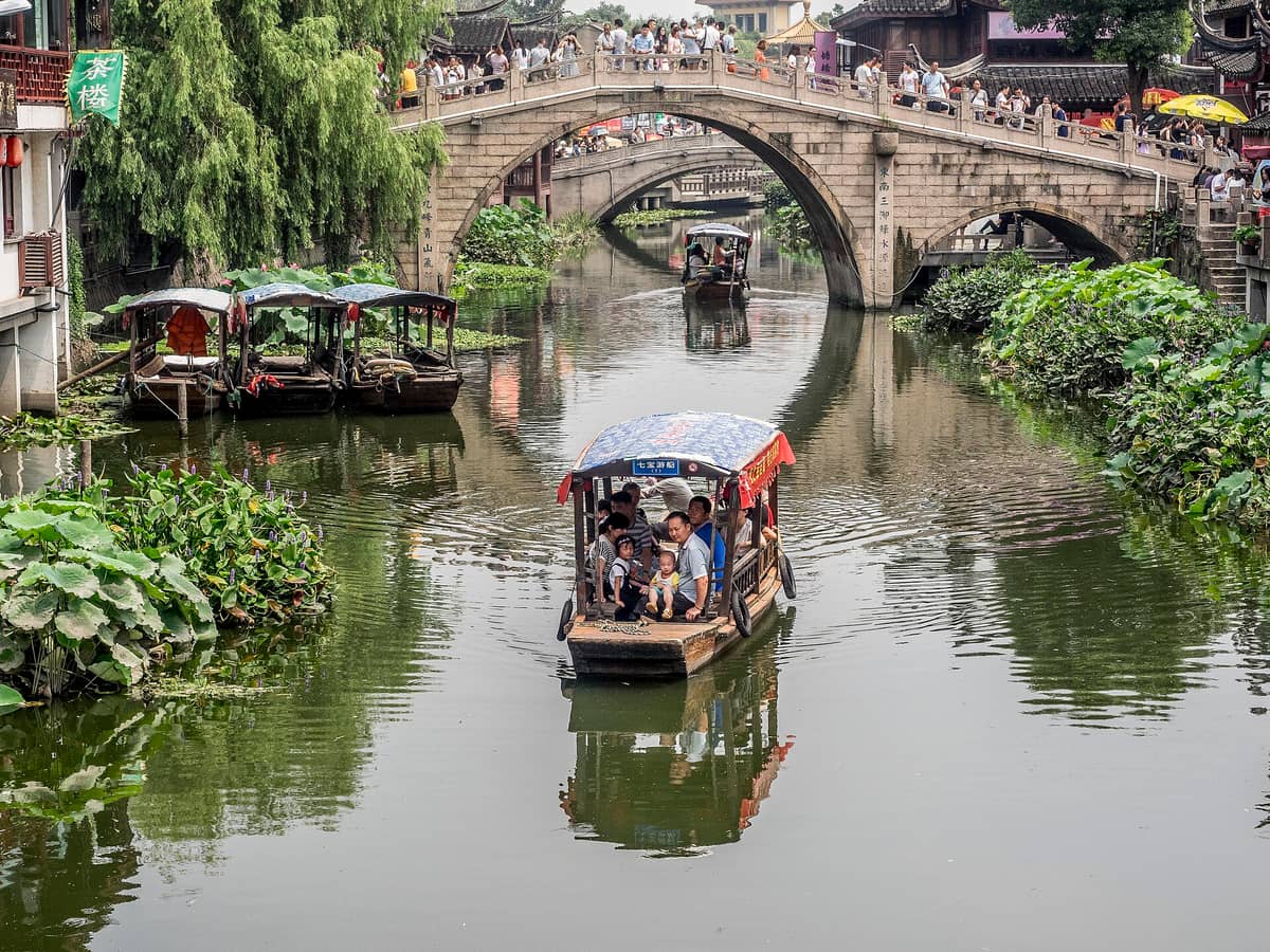 Ein Kanal in Qibao Ancient Town mit einer Steinbrücke, Holzbooten, üppigem Grün und historischen Uferhäusern voller Besucher.