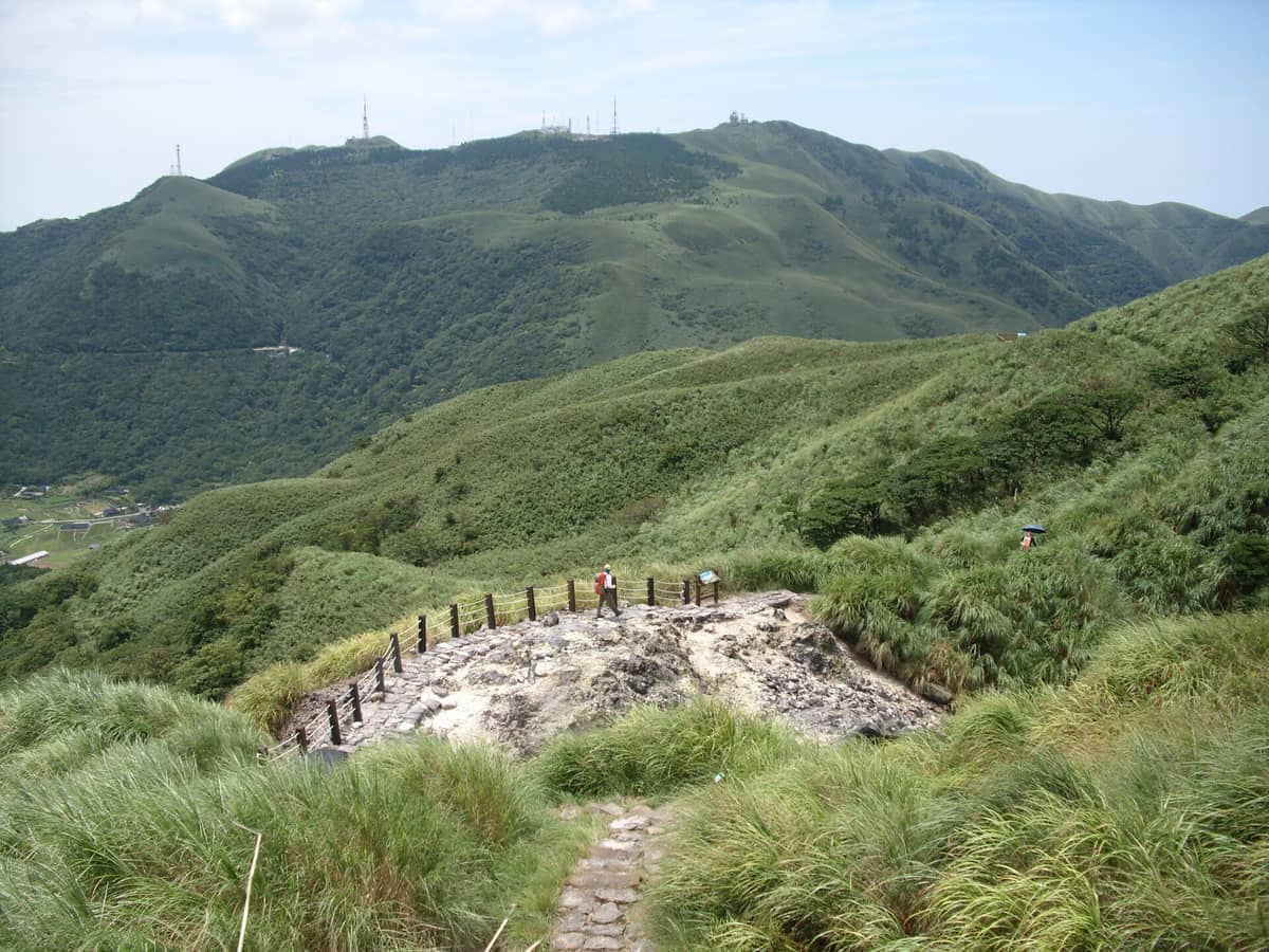 Gepflasterter Abschnitt des Qixing-Berg-Wanderwegs mit Wanderern, umgeben von üppigen grünen Hügeln und fernen Gipfeln unter klarem blauem Himmel.
