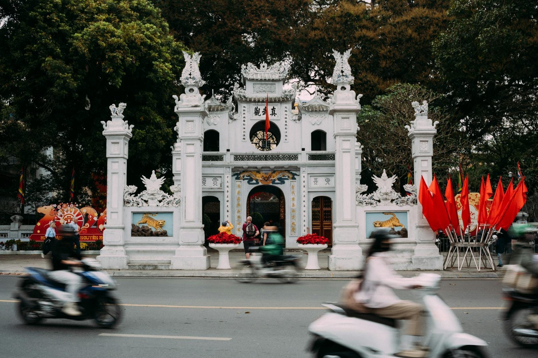 Front view of Quan Thanh Temple’s ornate white gateway with visitors and motorbikes passing by, set against leafy trees and vibrant red flags in Hanoi.