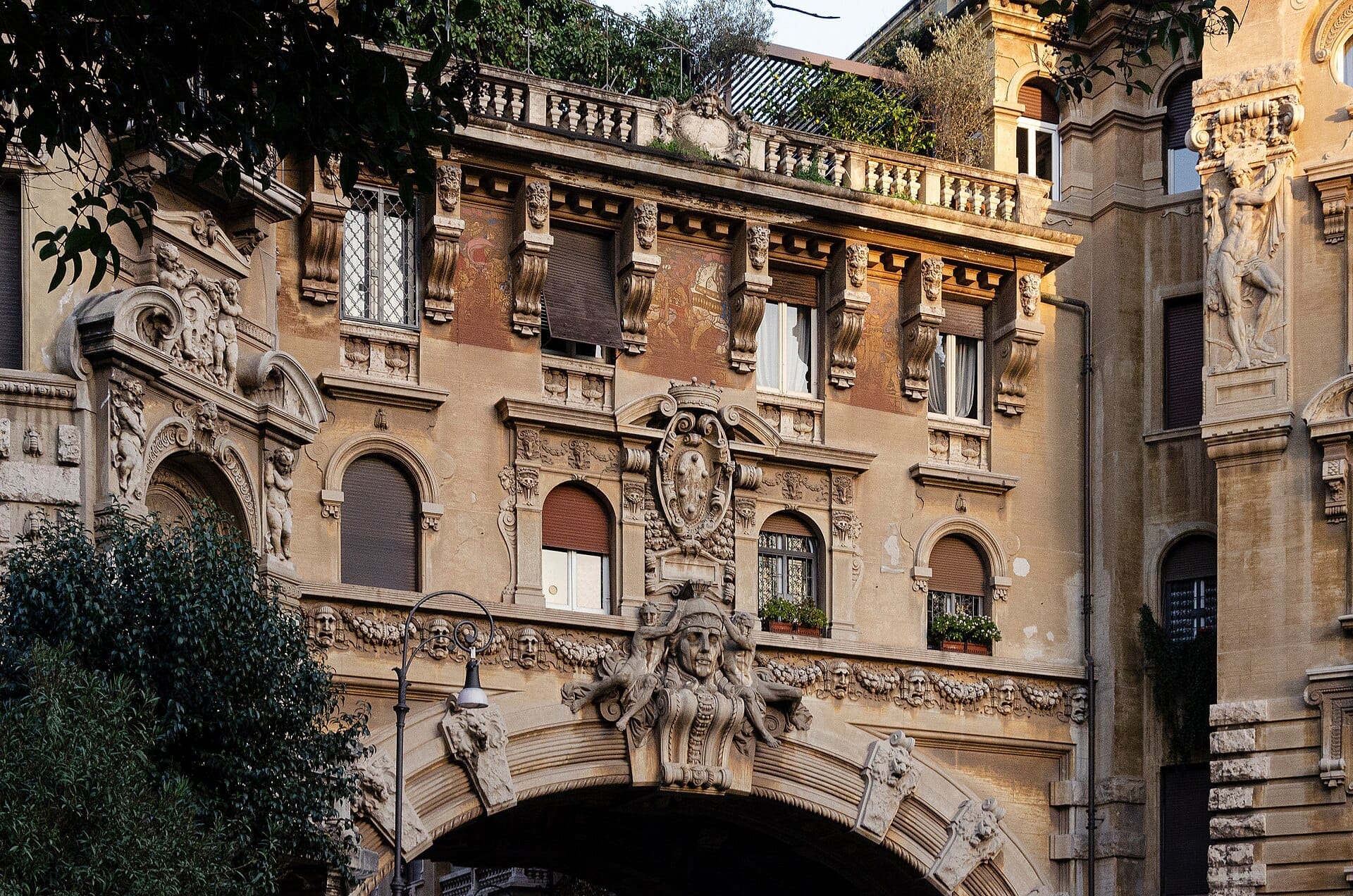 Close-up of Quartiere Coppedè’s ornate facades and decorative stone archway with intricate sculpted faces and greenery on balconies, under warm daylight.