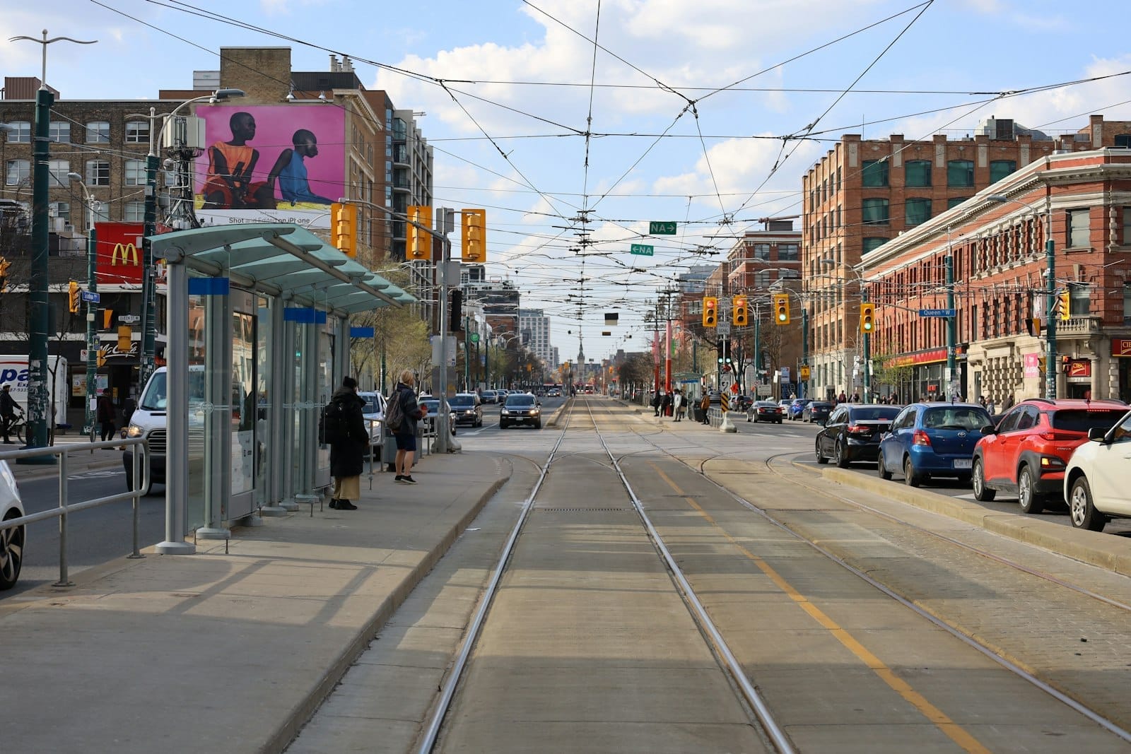 ถนน Queen Street West ในโตรอนโตช่วงกลางวัน มีรางรถราง ผู้คนรอที่ป้าย อาคารอิฐสวย และบรรยากาศเมืองที่มีชีวิตชีวา