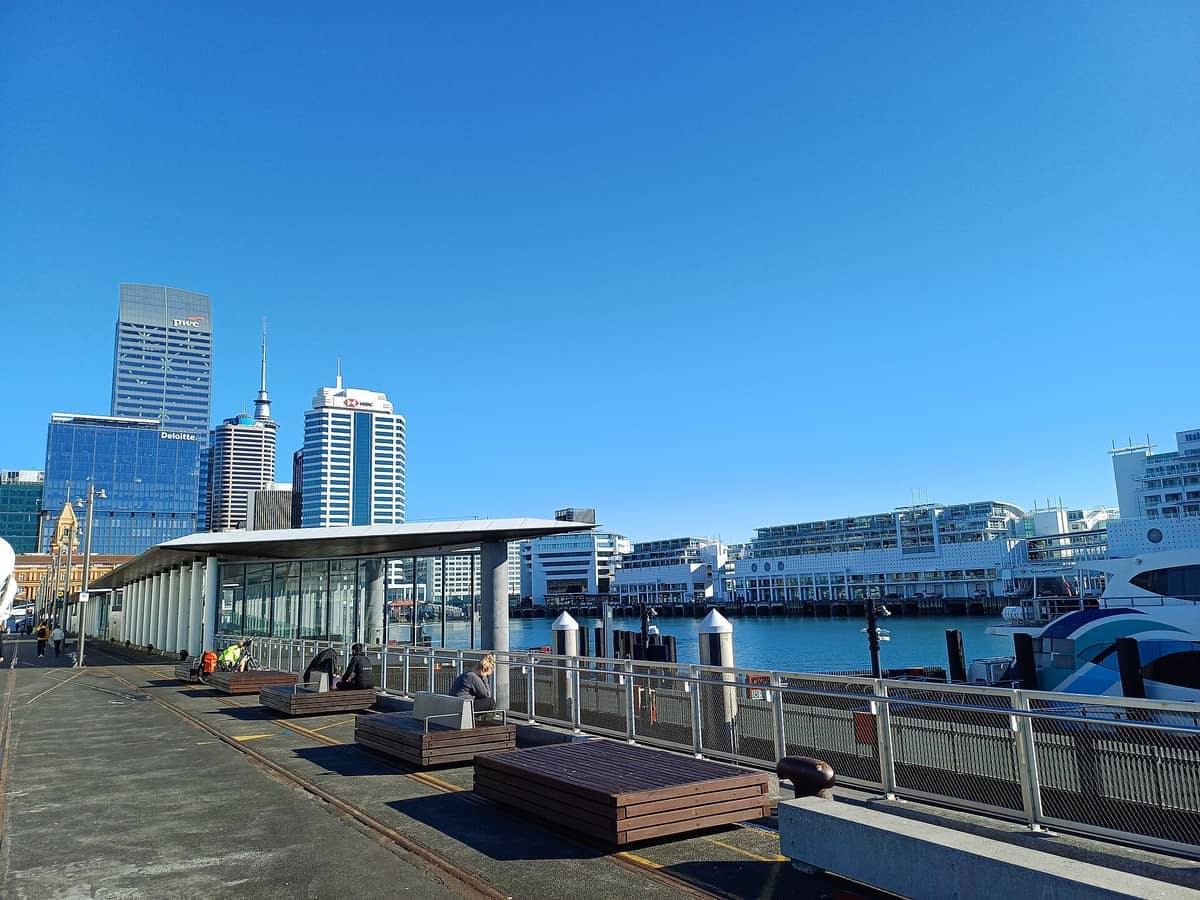 Queens Wharf in Auckland mit modernen Wolkenkratzern, Sitzbänken am Wasser, entspannenden Menschen und am Hafen angedockten Fähren unter einem strahlend blauen Himmel.