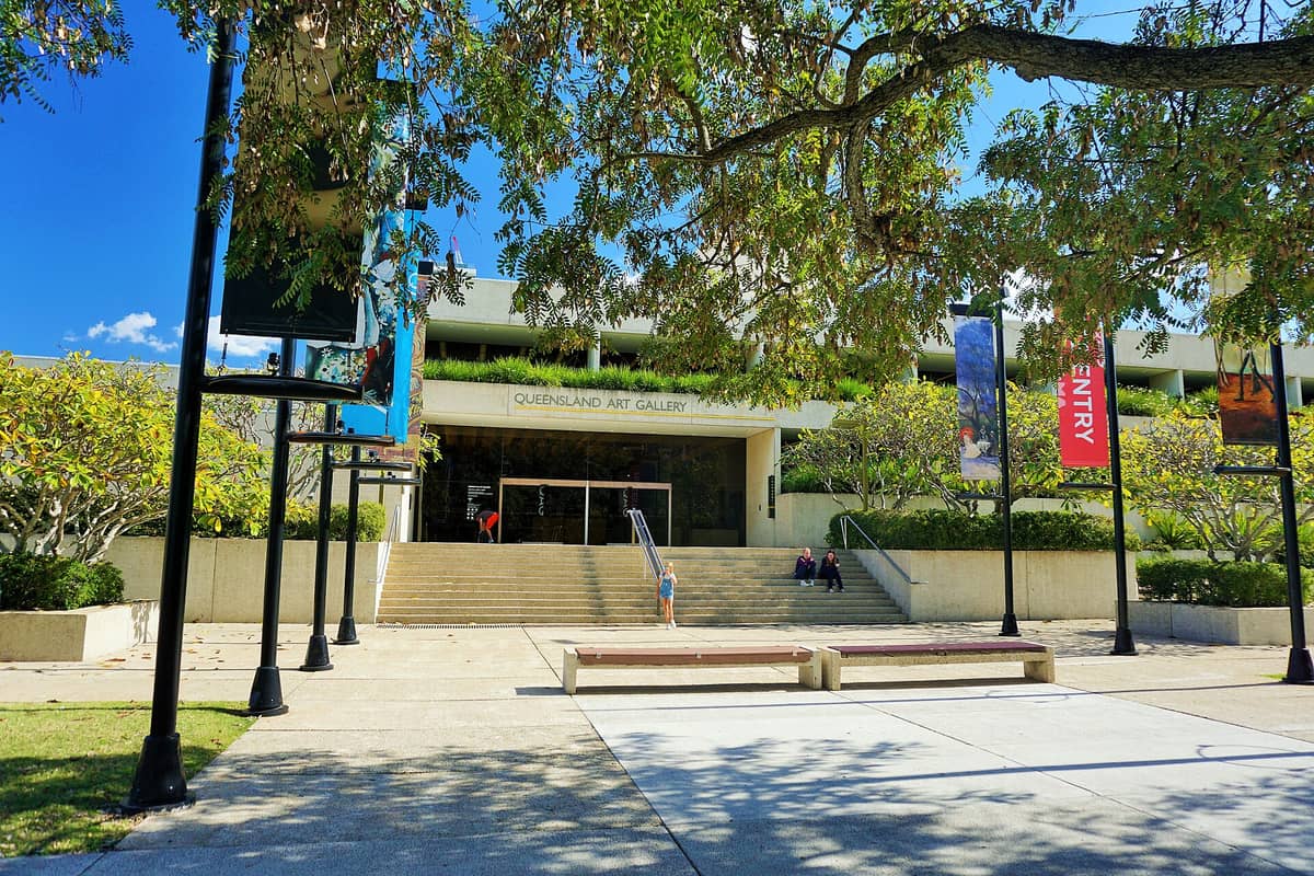 Wejście główne do Queensland Art Gallery w Brisbane — szerokie schody, zielone drzewa, szyldy galerii i błękitne niebo w tle.