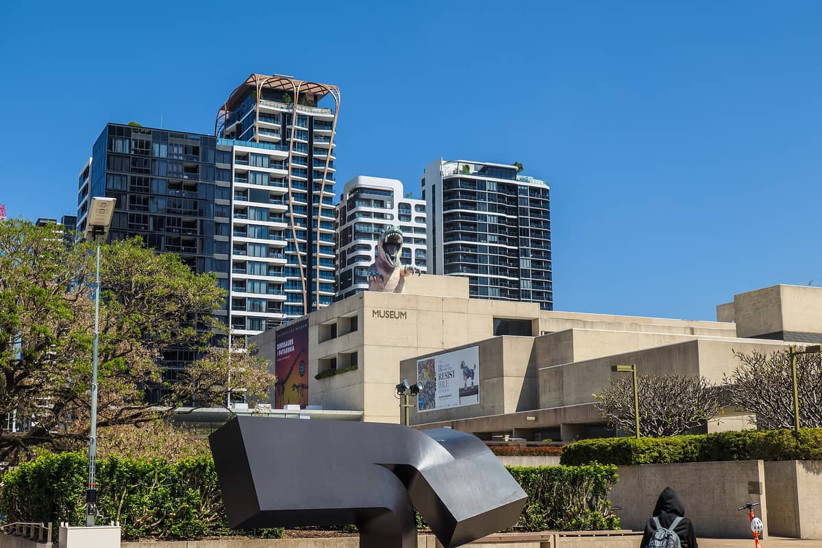 Fasada Queensland Museum Kurilpa z wyraźnymi napisami muzeum, banerami bieżących wystaw, nowoczesnymi budynkami miejskimi w tle i rzeźbą dinozaura na dachu.