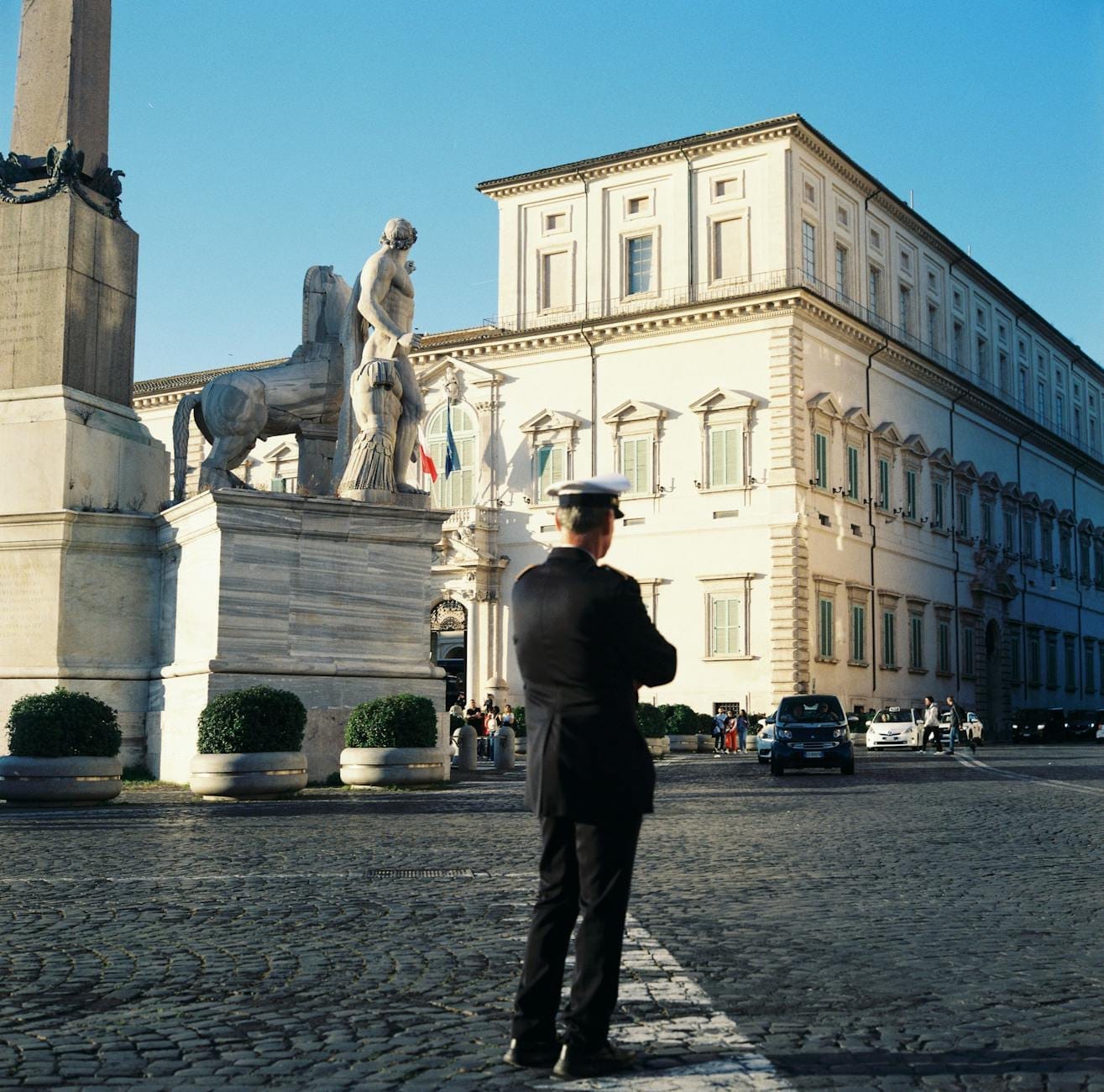 Quirinal Palace in Rome with a uniformed guard standing near the obelisk and statues, under a clear blue sky.