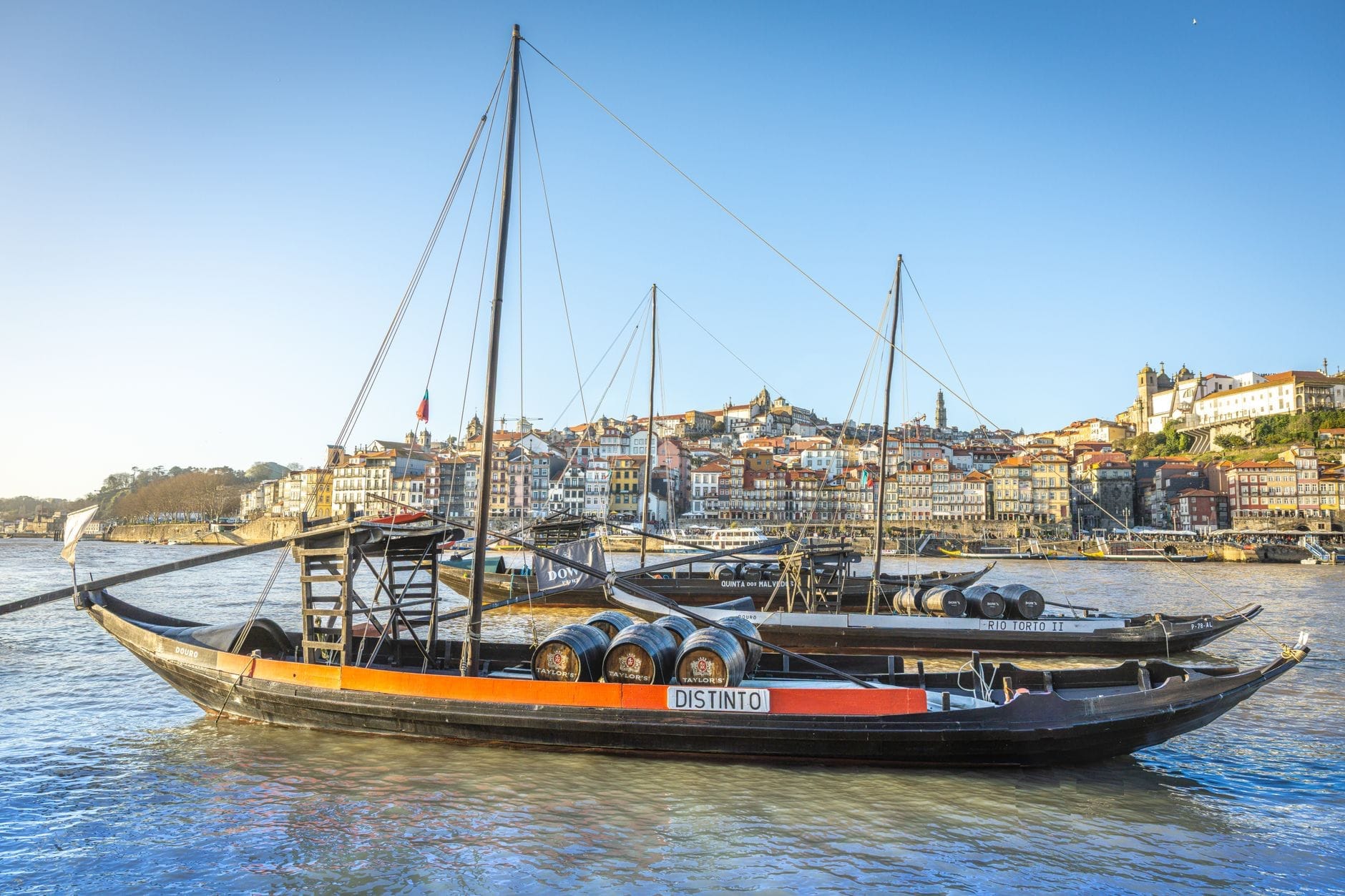 Tradizionali barcos rabelo con botti di vino sul fiume Douro, sullo sfondo la città di Porto e i colorati edifici sul lungofiume sotto un cielo azzurro.