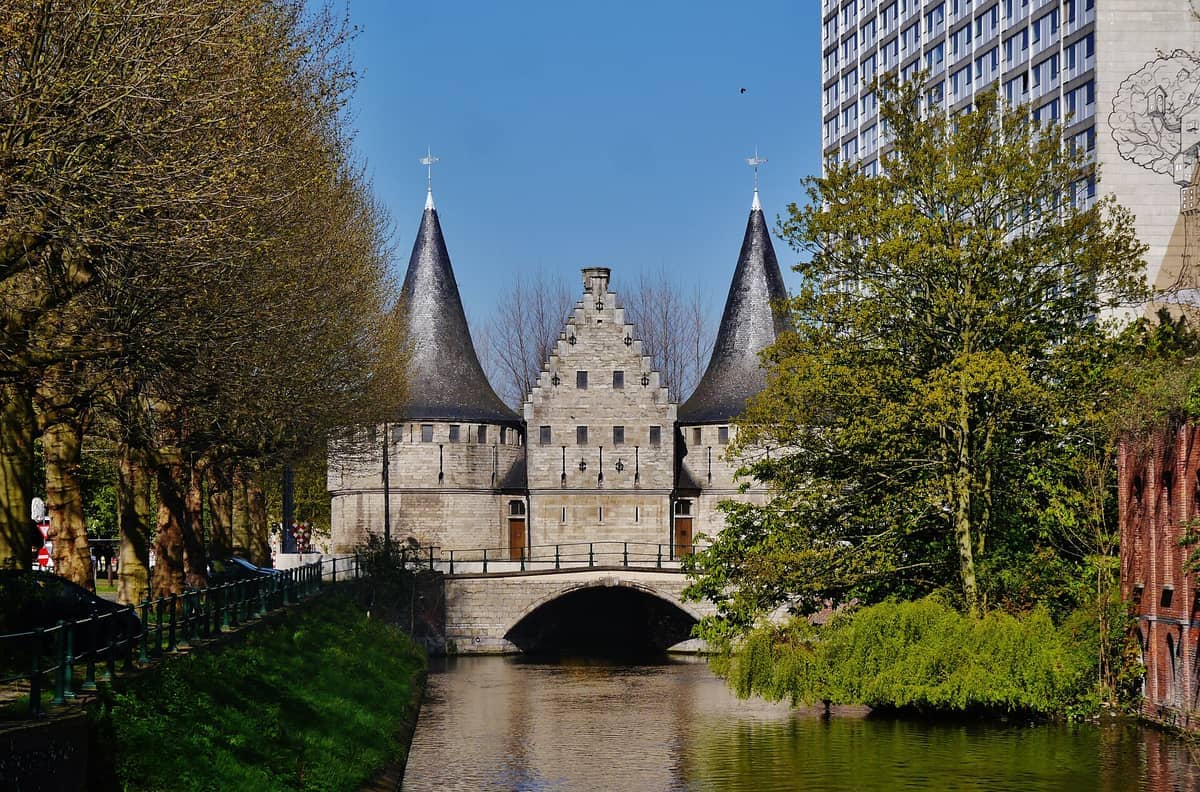 Rabot City Gate's twin medieval towers and archway span a serene canal in Ghent, bordered by trees and a modern high-rise nearby.