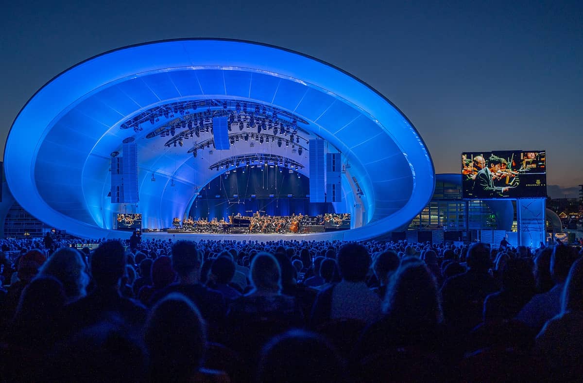Große Menschenmenge bei einem Nachtkonzert in der Rady Shell at Jacobs Park; die architektonisch markante, beleuchtete Shell-Struktur erstrahlt in blauem Licht.