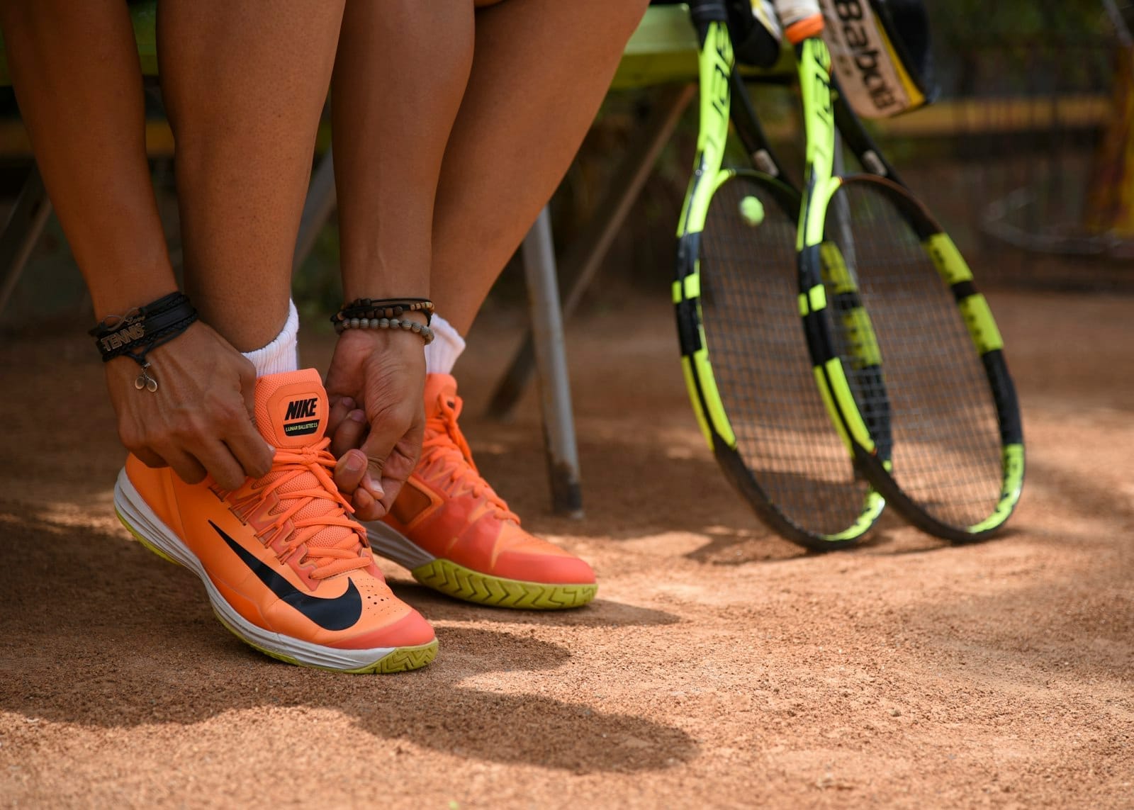 Primo piano di una persona che allaccia scarpe da tennis arancioni accanto a due racchette da tennis gialle su un campo in terra rossa al Rafa Nadal Museum Xperience.