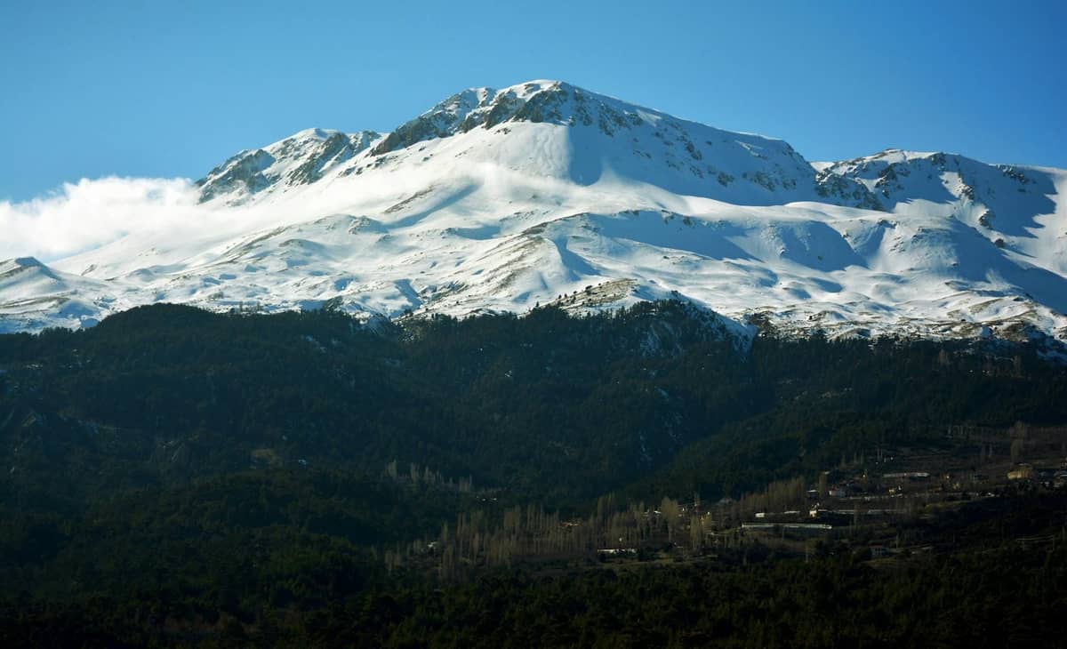 Snow-covered Taurus Mountains under a clear blue sky, forested lower slopes with a few scattered buildings.