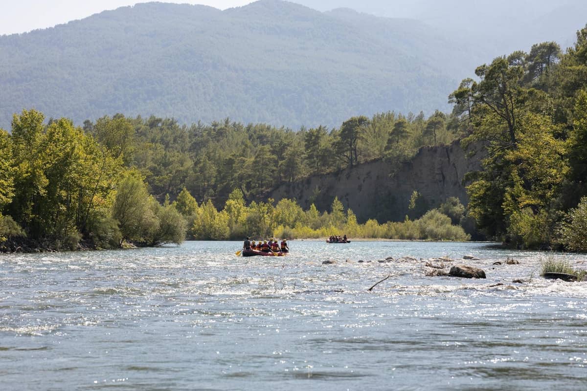 Wide view of rafts floating down a scenic river surrounded by forested banks and distant mountain landscape.