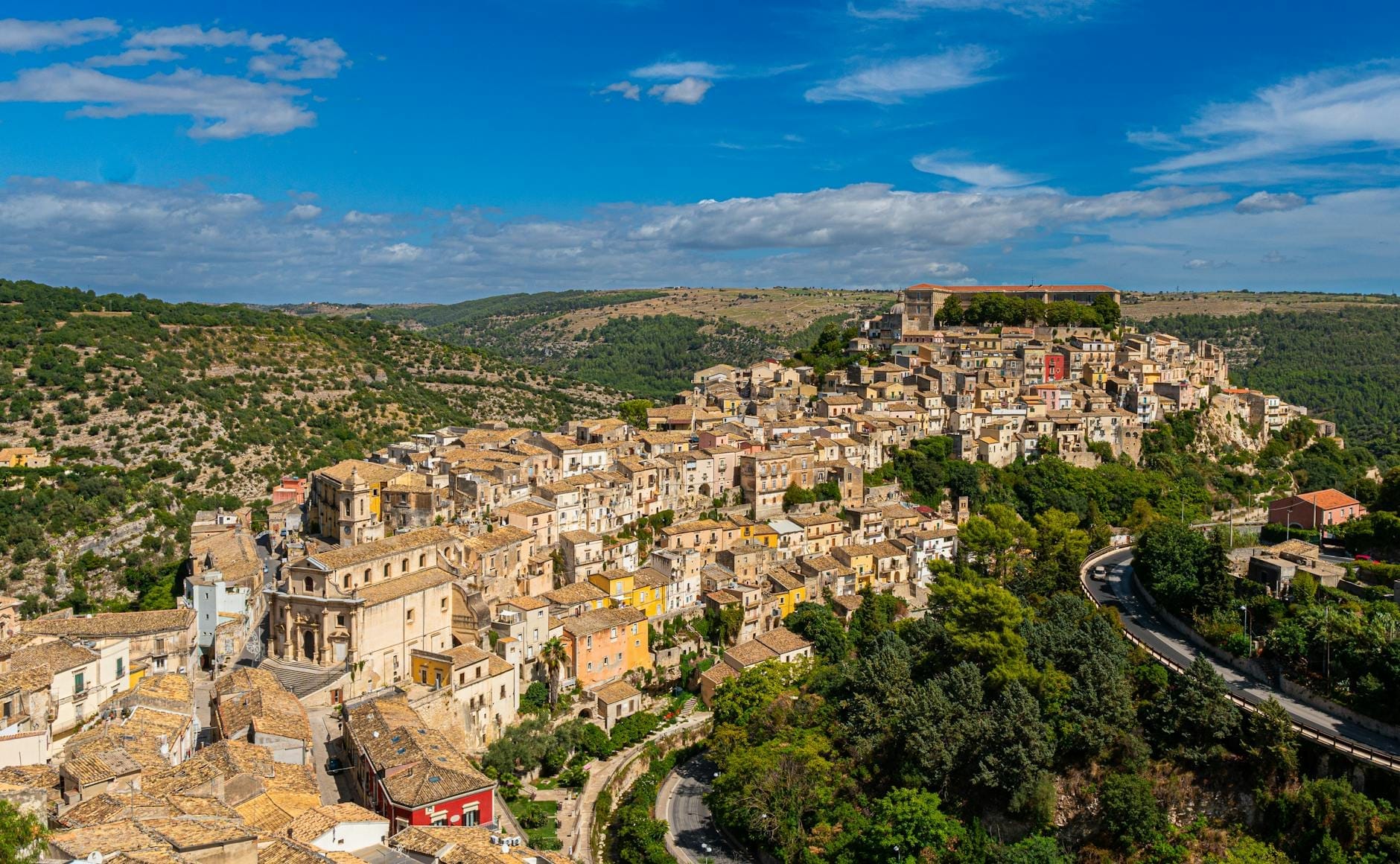 Weites Panorama von Ragusa Iblas historischen Barockgebäuden, die sich einen Hügel hinunterziehen, umgeben von sattgrünen Tälern unter einem strahlend blauen Himmel mit vereinzelten Wolken.