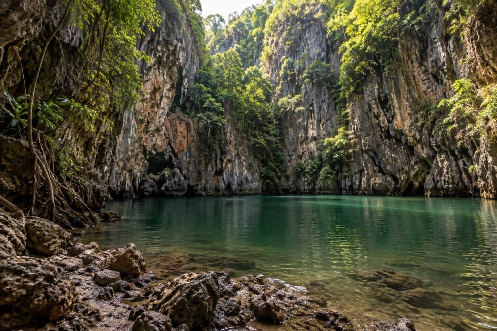 Railay Lagoon in Krabi: stilles türkisfarbenes Wasser, umschlossen von dschungelbewachsenen Kalksteinfelsen, vom felsigen Ufer aus. KI-Darstellung.