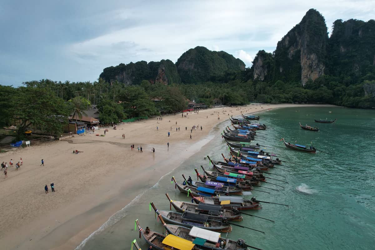 ภาพมุมสูงของชายหาดคาบสมุทรไร่เลย์ มีเรือหางยาวหลากสีจอดเรียงราย หาดทรายขาว นักท่องเที่ยวเดินเล่น และฉากหลังเป็นหน้าผาหินปูนเขียวขจีสูงชัน