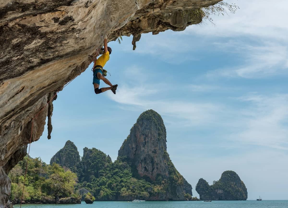 Escalador con camiseta amarilla afrontando un pronunciado saliente de roca caliza en Railay, con impresionantes acantilados costeros y aguas turquesas al fondo.