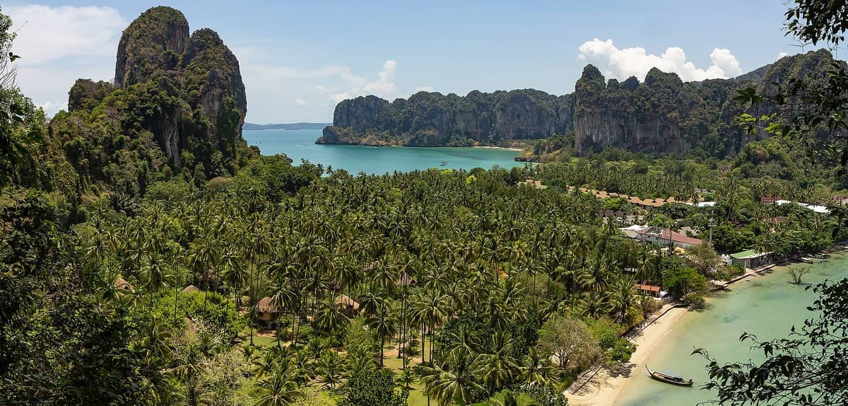 Panoramablick vom Railay Viewpoint mit üppigem Dschungel, Kalksteinfelsen, türkisfarbenem Wasser, kleinen Gebäuden und einem Sandstrand bei hellem Tageslicht.