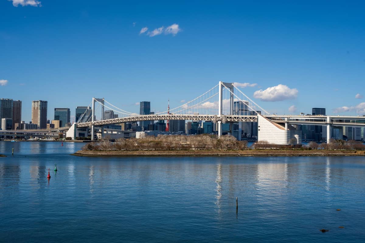 Daytime view of Tokyo’s Rainbow Bridge spanning Tokyo Bay, with the city skyline, water, and blue sky in the background.