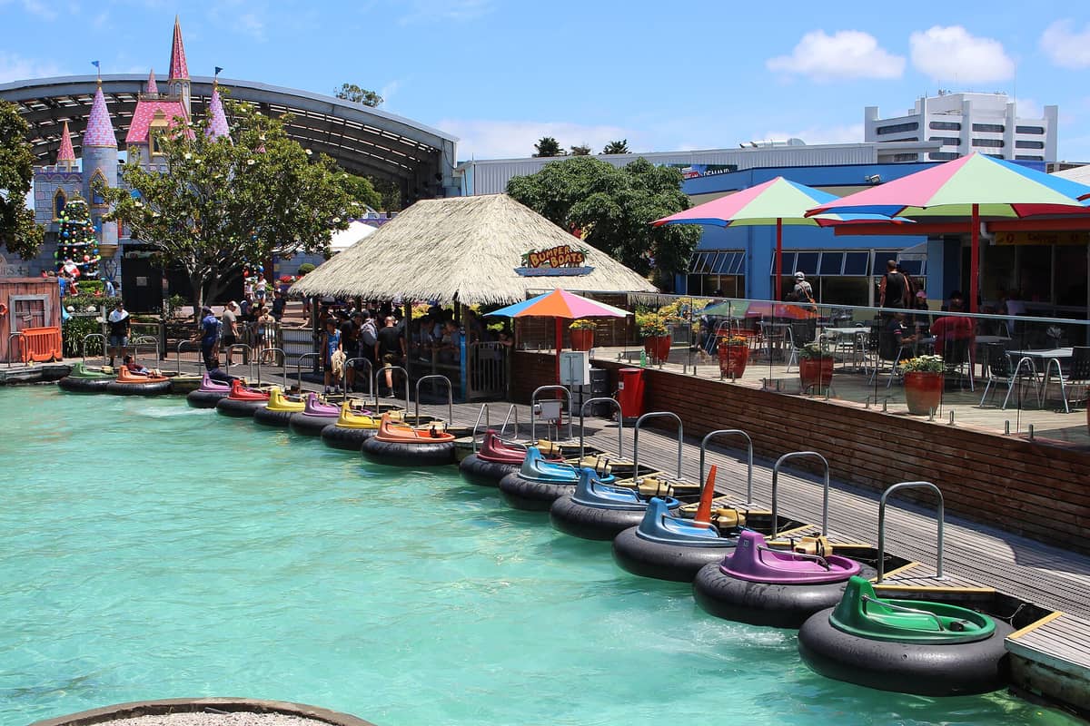 Die Bumper-Boats-Attraktion im Freizeitpark Rainbow's End in Auckland: bunte Boote auf einem Becken, wartende Besucher unter Sonnenschirmen, im Hintergrund das schlossartige Kidz-Kingdom-Areal.