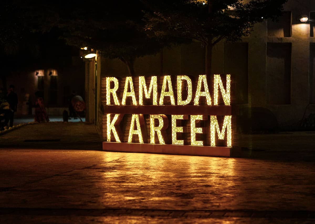 Beleuchtetes 'Ramadan Kareem'-Schild, das nachts in einem Innenhof leuchtet, mit dunklen Bäumen und einem gedämpften Hintergrund in einer urbanen Stadt im Nahen Osten.
