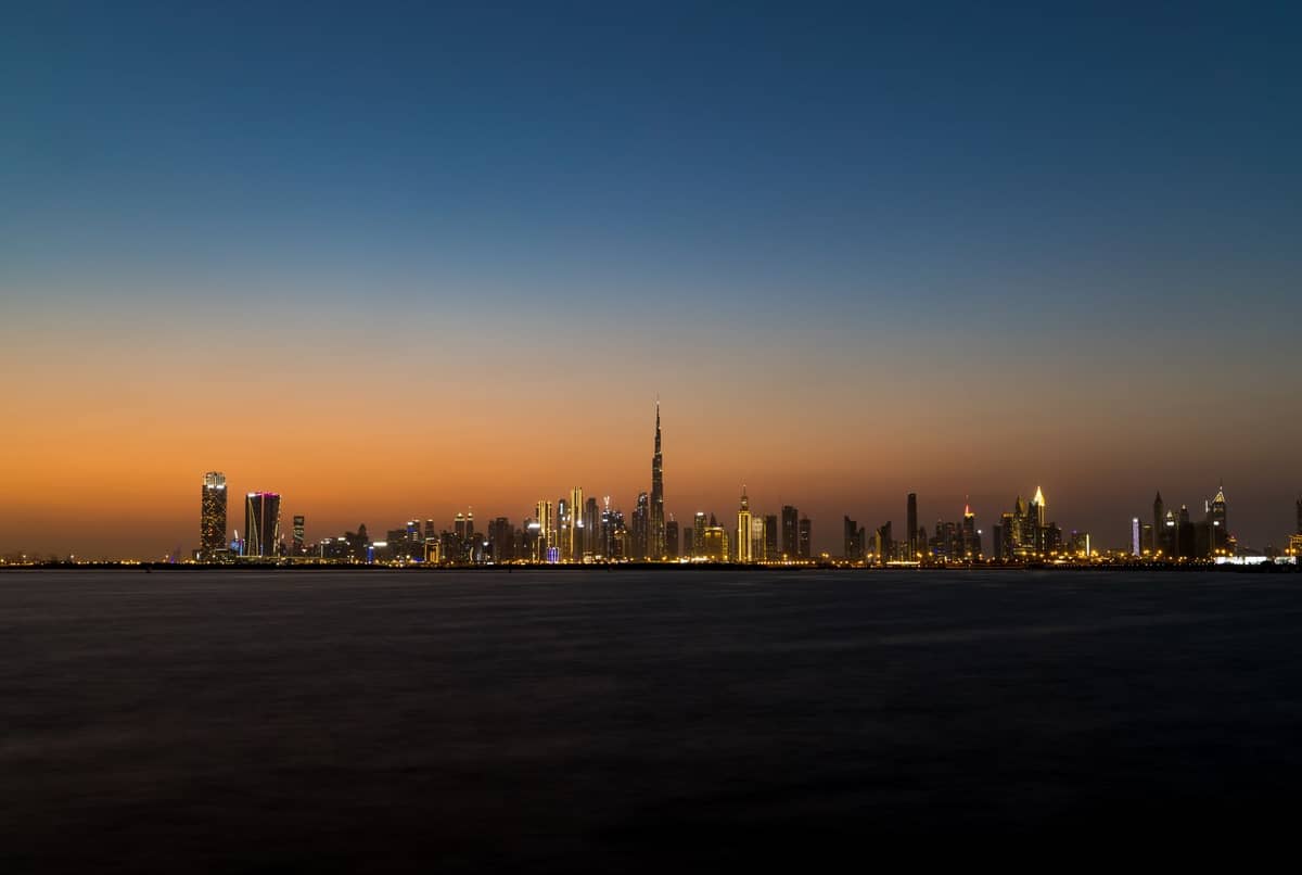 Weitwinkelpanorama der Dubai-Skyline in der Dämmerung mit dem markanten Burj Khalifa und glitzernden Stadtlichtern, gespiegelt im ruhigen Wasser unter einem Abendhimmel mit Farbverlauf.