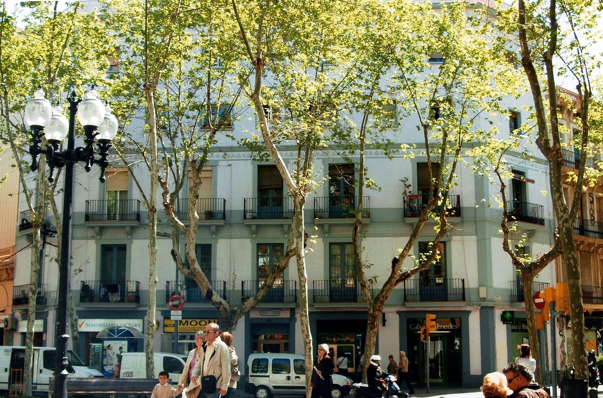 A tree-lined stretch of Rambla del Poblenou with classic low-rise buildings, leafy branches casting dappled sunlight, and locals strolling below.