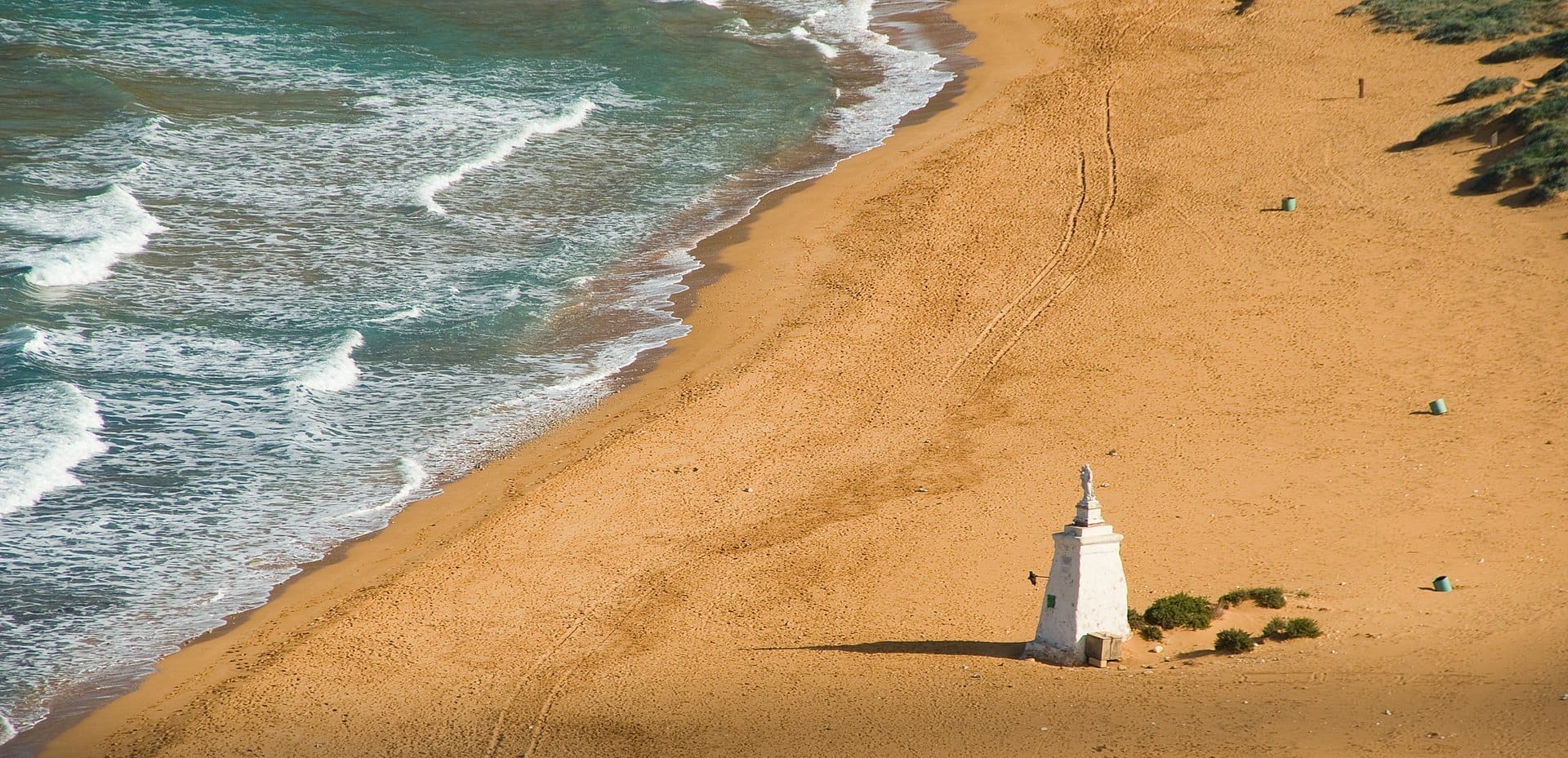 Vista aerea della riva di sabbia rossa di Ramla Bay, dolci onde blu, un solitario monumento con statua e vegetazione sparsa lungo l'ampia spiaggia.