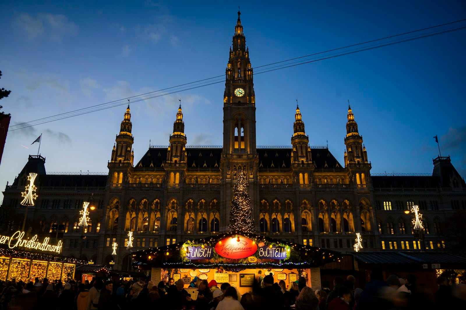 Das Wiener Rathaus bei Nacht mit beleuchteten neugotischen Türmen, festlichen Weihnachtsmarktlichtern und einer fröhlichen Winteratmosphäre im Vordergrund.
