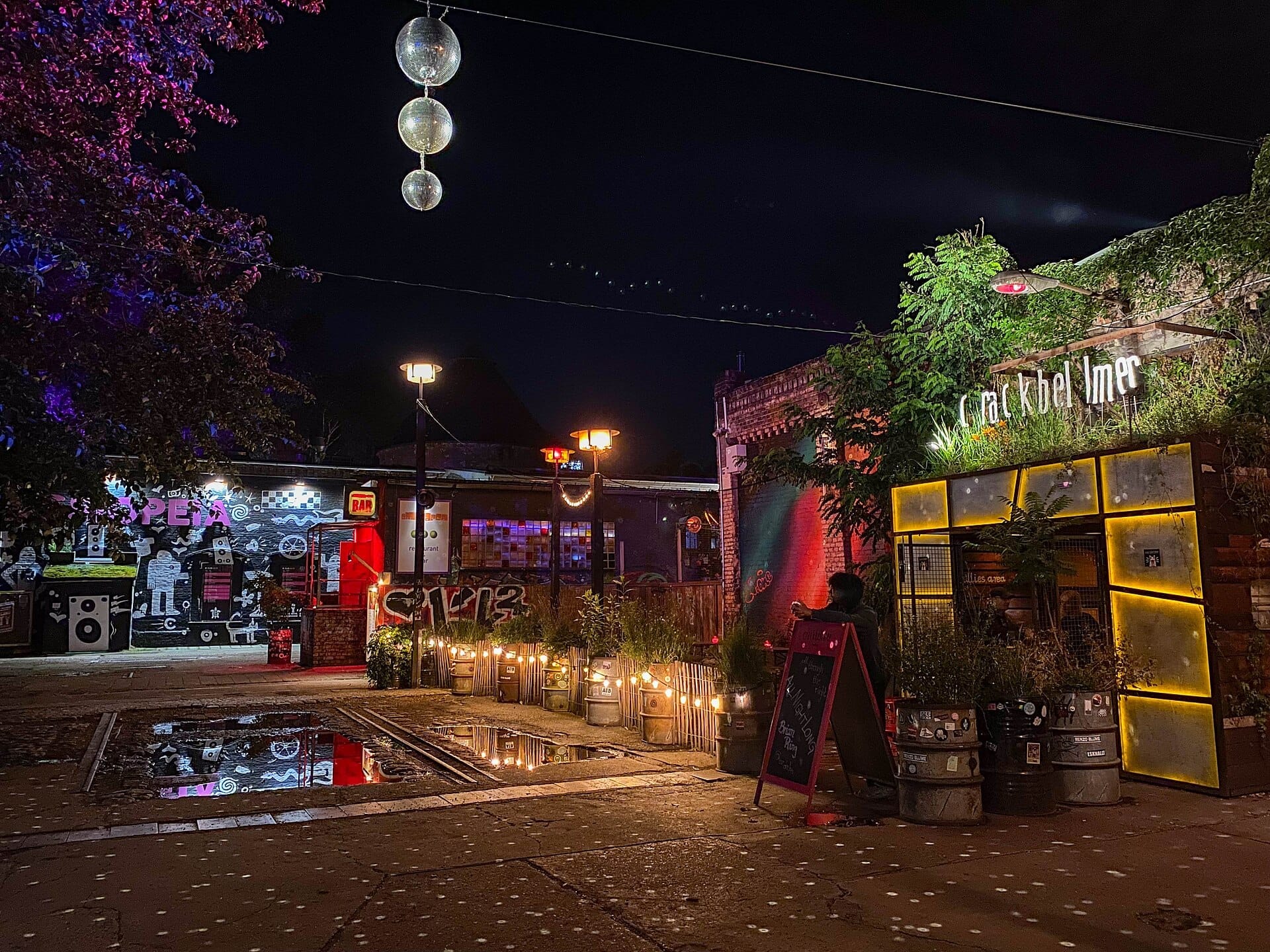 Vista notturna del RAW-Gelände a Berlino con bar illuminati, muri ricoperti di graffiti, fioriere a botte e palle da discoteca sospese in alto.