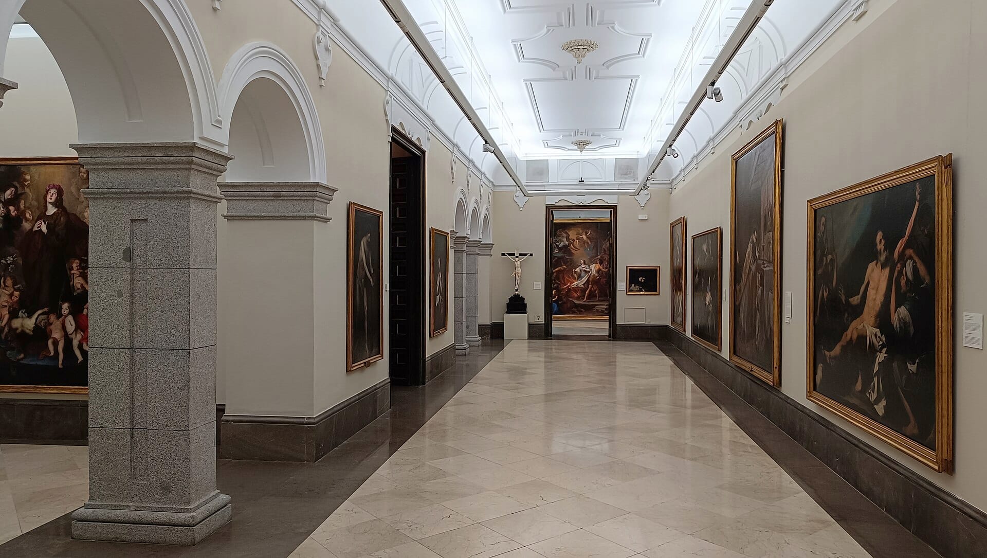 Ampia galleria con pavimento in marmo della Real Academia de Bellas Artes de San Fernando, fiancheggiata da grandi dipinti incorniciati e colonne classiche sotto una luce intensa.