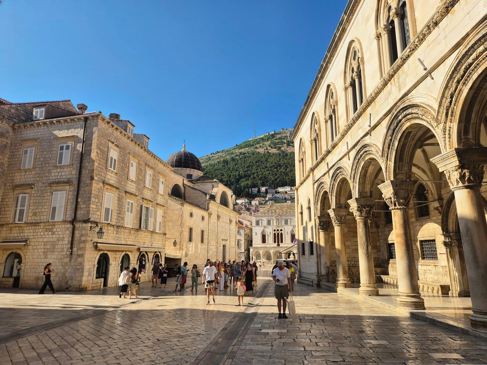 Vista amplia del Palacio del Rector en Dubrovnik con elegantes arcos, edificios históricos cercanos, personas explorando la soleada plaza del casco antiguo y las colinas de fondo.