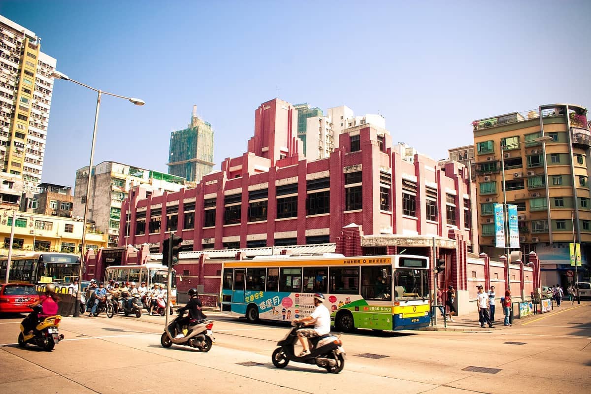 Der Red Market in Macau fällt mit seiner roten Backsteinfassade auf, umgeben von Stadtbussen, Rollern und Fußgängern an einem sonnigen Tag.