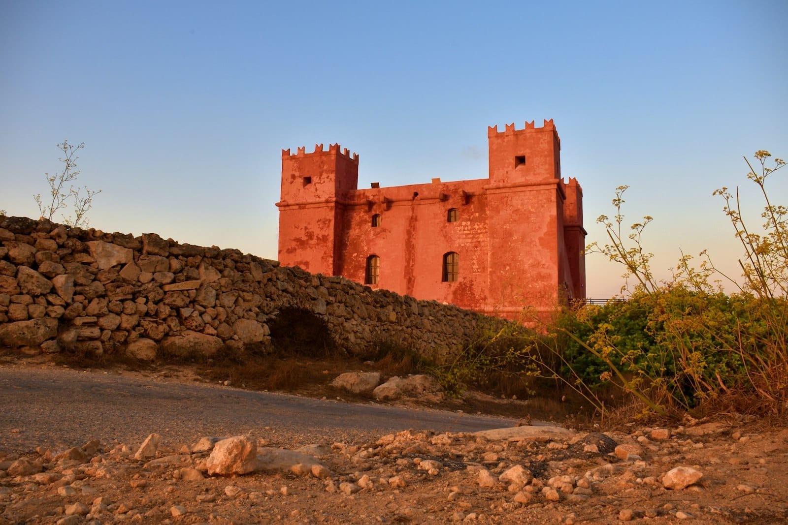 La Red Tower a Malta si erge sopra un muro a secco, la sua facciata rossa illuminata dalla calda luce della sera contro un cielo blu limpido.