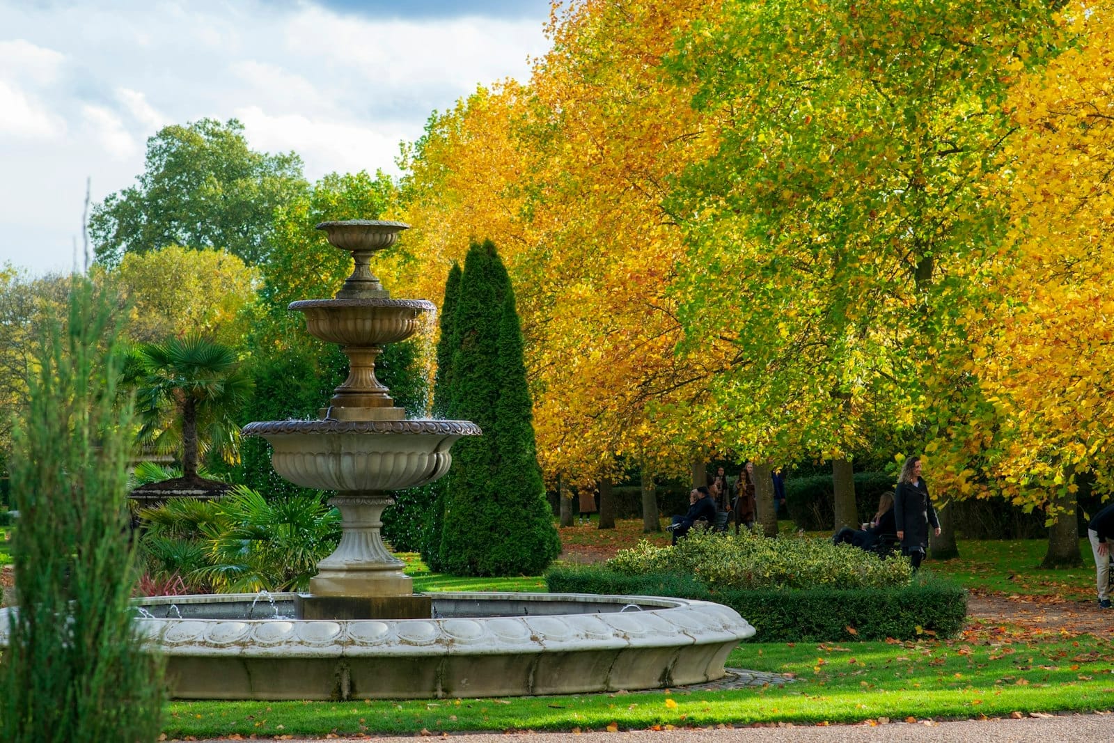 Fontana centrale in pietra circondata da rigogliosi prati e alberi autunnali dorati nel Regent's Park, con visitatori che passeggiano e si rilassano sulle panchine.
