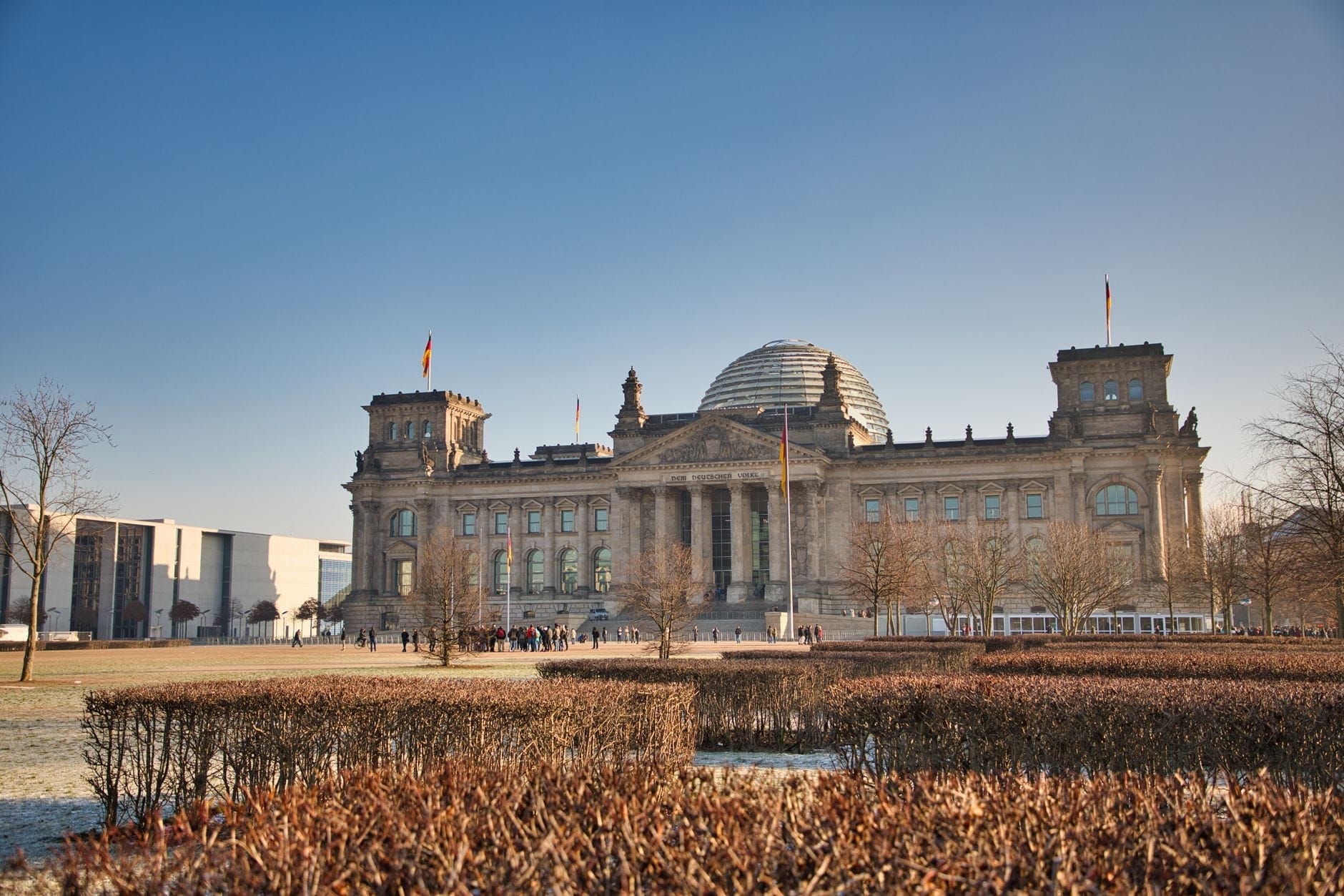 Vista panoramica del Reichstag di Berlino sotto un cielo azzurro limpido, con l'iconica cupola di vetro e le bandiere tedesche ben visibili, incorniciato dalla vegetazione invernale.