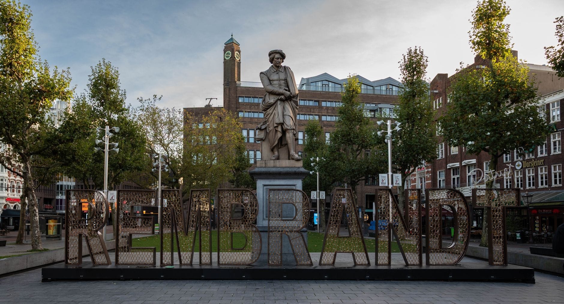 La estatua de Rembrandt preside el centro de Rembrandtplein en Ámsterdam, rodeada de árboles, edificios históricos y la plaza abierta bajo un cielo parcialmente nublado.