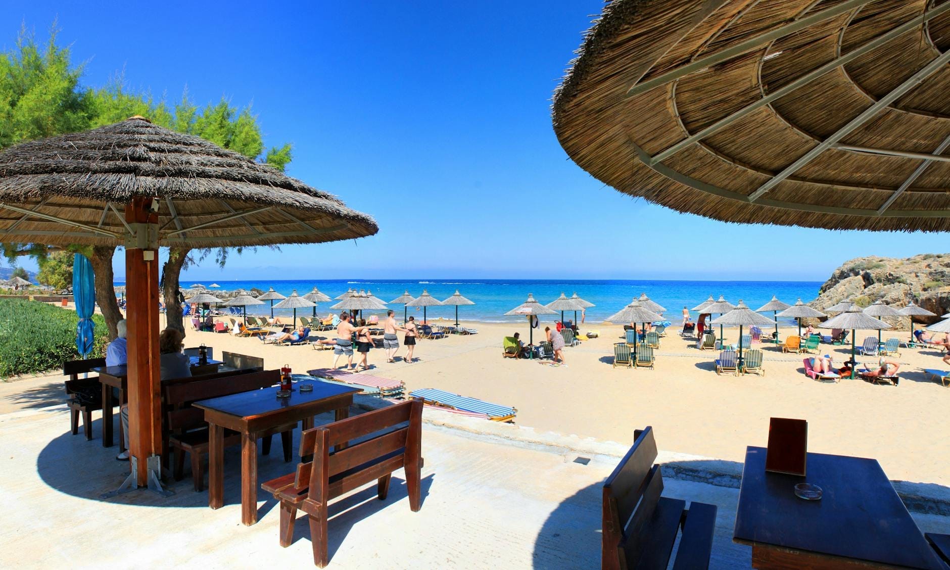 Vista da una taverna sul lungomare di Rethymno Beach con ombrelloni di paglia, lettini, persone che si rilassano e un mare blu cristallino sotto un cielo soleggiato.